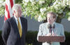 An eye-level, medium shot shows Bill Clinton, left, and President Kevin Payne , right, standing outdoors against a backdrop of blooming white trees. Clinton, wearing a navy blue suit and a patterned gold tie, stands with his hands behind his back, looking toward the man at the podium. The man at the podium, in a light gray suit and a red patterned tie, speaks into two microphones. An American flag is partially visible in the upper left corner.