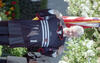 This medium, eye-level shot captures Bill Clinton smiling as he holds up a black D.C. United soccer jersey. He is dressed in a dark suit and stands in front of a blooming white tree, with a portion of the American flag visible in the background. The 1996-98 home jersey features white horizontal stripes across the chest, the Adidas logo, and the number "1". A small red and white D.C. United crest is visible on the left side.