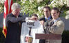n eye-level, medium shot shows Bill Clinton, left, and two unidentified player, right, standing outdoors against a backdrop of blooming white trees. Clinton, wearing a navy blue suit and patterned gold tie, smiles as he holds one side of a white D.C. United soccer jersey with the name "GORE" and the number "1" on the back.