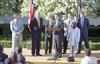 An eye-level, medium shot shows Bill Clinton, second from left, and several others on a stage outdoors at the White House. Clinton, in a navy suit and gold tie, stands with his hands behind his back. At the center, John Harkes in a dark suit speaks into microphones at a wooden podium.