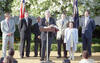 An eye-level, medium shot shows Bill Clinton speaking at a wooden podium during an outdoor event. Clinton, in a navy suit and patterned gold tie, holds a folded D.C. United soccer jersey while standing before two microphones.Surrounding him are several individuals, including three men in suits, a young boy, and a young girl in a light blue coat. They are gathered on a stage against a backdrop of flowering white trees, with the American flag and the Presidential Flag visible in the background.