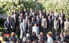 An eye-level, medium-wide shot captures Bill Clinton posing for a group photograph with the 1996 D.C. United team and staff. Clinton, wearing a navy suit and gold tie, stands on the left side of the second row.The large group, primarily composed of men in various suits and ties, is arranged in multiple rows on an outdoor stage. In the front row, a young boy and girl stand centrally. The group is set against a backdrop of lush green shrubbery and white flowering trees. 