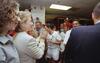 Hillary Clinton stands in profile facing right, wearing a cream-colored blazer and gold earrings, smiling as she claps. Chelsea Clinton stands slightly in front of her to the far left, also in profile, smiling with curly hair. They face soccer player Mia Hamm, who wears a white USWNT jersey with the number 9 and holds a water bottle. Several other teammates, coaches, and staff members are visible in the background smiling, while a man in a dark blazer is seen from the back on the right side.
