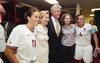 On the left, player number 11, Julie Foudy, smiles in her white jersey. Next to her, Hillary Clinton wears a cream-colored suit jacket, standing beside Bill Clinton, who wears a dark suit and gold tie. Chelsea Clinton stands to his right in a grey button-down shirt and black pants, with her arm around team captain Carla Overbeck, who wears her white number 4 jersey and captain's armband while holding a water bottle.