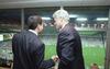 A photograph shows Bill Clinton shaking hands with a man wearing a pinstripe suit, viewed from behind as they stand inside a stadium suite looking out over a soccer field filled with spectators.