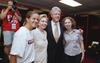 A photograph shows a Brandi Chastain wearing a white graphic t-shirt posing alongside Hillary Clinton, Bill Clinton, and Chelsea Clinton inside a locker room while a photographer stands in the background.