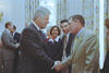 A medium shot shows former U.S. President Bill Clinton, wearing a dark pinstripe suit, shaking hands with an older man in a tan suit jacket and a patterned yellow tie in a brightly lit room. In the background, other individuals look on or converse, with patterned curtains hanging behind them.
