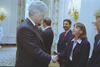 A medium shot captures former U.S. President Bill Clinton from the side profile as he smiles and shakes hands with a woman wearing a dark blazer over a light pink button-down shirt. A man with a mustache stands in a navy blazer behind her, while other guests and decorative gold sconces are visible in the background of the elegant room.