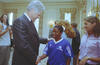 A medium shot shows former U.S. President Bill Clinton smiling as he shakes hands with a young Black girl wearing a blue Adidas sports jersey that reads "DC SCORES." He places his other hand gently on her shoulder. On the left, a photographer holds a camera, while another woman smiles on the right side of the frame, all set within a brightly lit, formal room with patterned curtains.