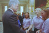 A medium shot captures former U.S. President Bill Clinton from a side profile as he smiles and shakes hands with a woman with short blonde hair. She is wearing a light pink short-sleeved button-down shirt, matching another smiling woman standing next to her. In the background, other guests stand in a formal, well-lit room decorated with patterned curtains and an ornate gold fireplace mantel.