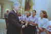 A medium shot captures former U.S. President Bill Clinton smiling and shaking hands with one of several women lined up in a well-lit room [1]. The women are smiling, with four wearing matching light pink short-sleeved button-down shirts and dark pants, while another wears a white button-down shirt. In the background, Vice President Al Gore stands on the left, and a large gold candelabra hangs on the ornate white wall.