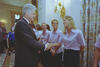 A medium shot captures former U.S. President Bill Clinton smiling as he shakes hands with a smiling, blonde Brandi Chastain. She stands in a line with other women wearing matching light pink short-sleeved button-down shirts and dark pants. In the background on the left, a young girl wears a blue sports jersey, while a gold wall sconce and patterned floral curtains decorate the elegant room.