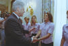 A medium shot captures former U.S. President Bill Clinton smiling and shaking hands with a smiling woman with curly hair who stands in a line. She and the other women behind her wear matching light pink short-sleeved button-down shirts. In the background, a person with a video camera films the interaction, set against a white wall with a gold sconce and floral curtains.