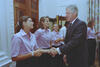 A medium shot captures former U.S. President Bill Clinton smiling broadly as he shakes hands with a woman with her hair pulled back into a ponytail. She and the other women standing beside her are wearing matching light pink short-sleeved button-down shirts and dark pants. The interaction takes place near a large, ornate wooden door frame within a well-lit room featuring floral-patterned curtains in the background.
