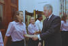 A medium shot captures former U.S. President Bill Clinton smiling as he shakes hands with a woman with shoulder-length brown hair. She and another smiling woman behind her are wearing matching light pink short-sleeved button-down shirts and dark pants. In the background, a woman in a white shirt looks on near a large wooden door, and floral-patterned curtains hang on the right side of the room.