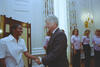 A medium shot captures former U.S. President Bill Clinton smiling as he shakes hands with a smiling Black woman wearing a white short-sleeved button-down shirt. On the right, three other women wearing matching light pink shirts and dark pants smile and watch the interaction. The group stands in an elegant room featuring tall windows with floral-patterned curtains and an ornate gold light fixture mounted on the white paneled wall.