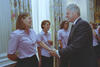 A medium shot captures former U.S. President Bill Clinton smiling as he shakes hands with a laughing, brown-haired woman. She is standing at the front of a line of women who are all smiling and wearing matching light pink short-sleeved button-down shirts with dark pants. In the background, tall windows are framed by floral-patterned curtains, and an ornate gold candelabra stands on a mantel to the far right.