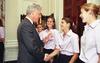 A medium shot captures former U.S. President Bill Clinton smiling as he shakes hands with a laughing woman with her hair in a ponytail. He holds his other hand up in a gentle gesturing wave. She and the other smiling women in line with her wear matching light pink short-sleeved button-down shirts and dark pants. The interaction occurs in an elegant room near a large dark wood door and white paneled walls.