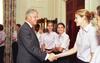 A medium shot captures former U.S. President Bill Clinton smiling as he shakes hands with a smiling blonde woman with her hair secured in a bun. She stands at the front of a line of women who are wearing matching light pink short-sleeved button-down shirts and dark pants. The greeting occurs near a large dark wood door frame, with floral-patterned curtains visible in the background on the far left.