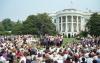 A large crowd of people sits on folding chairs and on the lawn of the White House South Portico, facing a small raised stage. On the stage, President Bill Clinton, First Lady Hillary Clinton, and several other officials and athletes stand at a podium. The iconic white, columned facade of the White House rises in the background under a clear sky, decorated with numerous American flags along its balcony railings. Large green trees flank the lawn, framing the gathering.