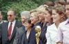 A close-up photograph captures former President Bill Clinton and First Lady Hillary Clinton standing outside with a group of people, including members of the U.S. Women’s National Soccer Team.Hillary Clinton stands near the center, smiling in profile. To her left is former President Bill Clinton, also smiling. A gold and silver World Cup trophy stands prominently in the foreground. Several smiling women and a man in a suit and sunglasses stand alongside them against a background of green trees.