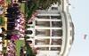 A wide shot captures the South Portico of the White House, where former U.S. President Bill Clinton and First Lady Hillary Clinton stand alongside Vice President Al Gore and Second Lady Tipper Gore on a small stage. They are gathered with a large group of women wearing matching light pink shirts and dark pants, and a young girl in a blue "DC SCORES" sports jersey speaks into microphones at a wooden podium. A crowd sits on the South Lawn grass in front of the stage, flanked by multiple American flags.