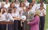 An outdoor ceremony at the White House shows Al Gore holding a white Nike soccer ball in the center. He wears a grey suit jacket and a red patterned tie. To his right, Tipper Gore stands in profile, wearing a magenta blazer and smiling toward the team. On the left side of the frame, several members of a women's soccer team shake hands near a wooden podium with two microphones. The team members wear matching light purple button-down shirts and dark pants, and many are smiling and clapping in the background.