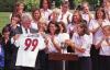 An outdoor White House ceremony features Bill Clinton holding up a white soccer jersey with the name "CLINTON" and the number "99" printed on the back. He stands on the left in a dark pinstripe suit. Two female soccer players stand next to him behind a wooden podium, smiling and clapping. A large group of team members, dressed in matching light purple button-down shirts, are gathered closely behind them, clapping and cheering in bright daylight.