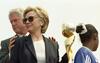 A close-up shot captures First Lady Hillary Clinton looking off-camera while wearing a black blazer, a white top, sunglasses, and gold earrings. Former U.S. President Bill Clinton stands partially behind her, placing his hand on her shoulder. On the right, the back of a young Black girl's head is visible in a blue Adidas sports jersey with a white scrunchie in her hair, alongside a portion of a gold trophy.