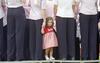 A medium shot from behind focuses on a young girl in a red and white gingham dress standing on a stage, holding the hand of a woman in a light pink shirt and dark pants. The girl looks toward the camera with a serious expression, framed by the legs and backs of multiple adults standing in line wearing matching pink or white shirts and dark trousers.