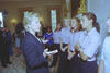 A medium shot captures First Lady Hillary Clinton from a side profile as she gestures with open hands while speaking to a group of listening women. The women stand closely together in a line, wearing matching light pink short-sleeved button-down shirts and dark pants. In the background, a young girl in a blue sports jersey stands nearby, while multiple news cameras and media personnel film the gathering in a brightly lit room decorated with floral curtains and an ornate gold fireplace mantel.