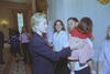 A medium shot captures First Lady Hillary Clinton smiling as she interacts with a group of women in a formal White House room. She places her hand on the back of a young girl wearing a red and white gingham dress, who is being held by a smiling woman in a white short-sleeved shirt. Other smiling women in matching light pink shirts look on, while a young girl in a blue sports jersey stands further back in the hallway.