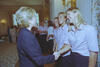A medium shot captures First Lady Hillary Clinton from a side profile as she shakes hands with a laughing blonde woman standing at the front of a line. She and the other women behind her wear matching light pink short-sleeved button-down shirts and dark pants. The greeting takes place in a brightly lit room next to a white wall with an ornate gold sconce and floral-patterned curtains on the far right.