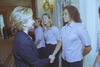 A medium shot captures First Lady Hillary Clinton from a side profile as she smiles and shakes hands with a smiling woman with curly hair. Two other women stand behind her in line, all wearing matching light pink short-sleeved button-down shirts and dark pants. The interaction occurs near a white wall decorated with an ornate gold sconce and floral-patterned curtains on the right.