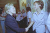 A medium shot captures First Lady Hillary Clinton from a side profile as she smiles and shakes hands with a smiling woman with short brown hair. Other women stand in a line behind her wearing matching light pink short-sleeved button-down shirts, with one taking a picture with a small camera. On the far left, a man wearing a striped button-down shirt and jeans stands next to a video camera, with floral-patterned curtains framing the wall on the right.