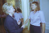 A medium shot captures First Lady Hillary Clinton from a side profile as she laughs and shakes hands with a smiling woman with shoulder-length brown hair. The woman wears a light pink short-sleeved button-down shirt and dark pants, matching the other women visible in the background. They stand in an elegant room next to white walls and tall windows draped with floral-patterned curtains.