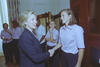 A medium shot captures First Lady Hillary Clinton from a side profile as she smiles and shakes hands with a smiling brunette woman. Behind them, another woman with her blonde hair in a ponytail stands with her hand on her chin watching the greeting. A Black woman in a white button-down shirt stands further back near a dark wood door, while two other women in light pink shirts talk on the far left next to floral-patterned curtains.