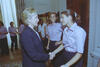 A medium shot captures First Lady Hillary Clinton from a side profile as she smiles and shakes hands with a smiling woman with her hair pulled back in a ponytail. Behind her, another woman with shoulder-length brown hair smiles and stands with her hand on her hip. In the background, a Black woman in a white button-down shirt and several other women wearing matching light pink shirts stand near a large wood door frame and floral-patterned curtains.