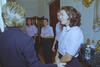 A medium shot captures First Lady Hillary Clinton from behind as she greets a laughing woman with shoulder-length brunette hair. Behind her, three other women smile and look on, all wearing matching light pink short-sleeved button-down shirts and dark pants. The interaction occurs in an elegant room next to a marble tabletop holding a bottle of water, with an ornate gold wall sconce mounted on the right.