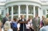 An outdoor White House ceremony features Hillary Clinton speaking at a wooden podium in the center, raising both hands with open palms. She wears a dark pantsuit. To her left stands Bill Clinton in a dark grey suit and a striped tie, looking toward her. To her right stands Al Gore in a light grey suit and a patterned red tie, smiling with his left hand in his pocket. A young boy in a blue and black uniform stands next to Bill Clinton. Members of the US Women;s World Cup team stand behind them.