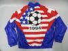 The back view of a vintage tracksuit jacket is laid flat, featuring a bold red, white, and blue graphic design celebrating the 1994 World Cup. The center panel displays a large black and white soccer ball set against a background mimicking the American flag, with white stars on a blue field to the left and horizontal red and white stripes across the rest of the panel. The year "1994" is printed in large black numbers directly beneath the soccer ball. The jacket has solid blue sleeves.