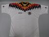 A white short-sleeved football jersey featuring a subtle, repeating geometric jacquard pattern woven into the fabric is laid flat against a light grey background. A bold graphic chevron pattern in the colors of the German flag—yellow, red, and black—stretches across the chest, shoulders, and collar area in a pixelated diamond gradient design. The jersey has a black collar trimmed with yellow and red stripes. The black Adidas wordmark is printed on the right chest, and the circular "Deutscher Fussball-Bund" 