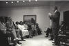 Black and white photograph. President Clinton stands while addressing a sitting audience of member of the Congressional Black Caucus.