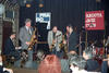 President Clinton stands and plays a brass saxaphone with two other people also standing and playing saxaphones on a stage. A man sits playing the piano behind President Clinton. Miphone stands and a large black curtain can also be seen in the background.