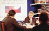 President Clinton is sitting across a table of others. He holds his left hand out in a gesture while in the middle of speaking. A large poster is on the wall in the background featuring the American and Czech Republic Flag. 