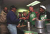 President Clinton and First Lady Hillary Rodham Clinton serve a Thanksgiving meal. The President and First Lady wear aprons and stand next to two other kitchen volunteers. They serve two men standing in line. 