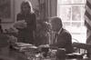 Black and white photograph. President Clinton meets in the White House Oval Office with the Director of the Office of Scheduling, Stephanie Streett. President Clinton sits behind the resolute desk. Stephanie Streett stands holding a stack of papers.
