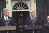 President Clinton and Prime Minister John Major stand behind podiums in front of a house. 