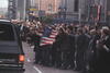 A crowd stands in the street, waving an United States flag at a black Chevorrolet Suburban. People in the crowd wave and clap.