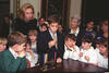 First Lady Hillary Rodham Clinton stands behind a group of children. A boy in a blue jacket lights a menorah. A menorah sits on a table in front of the children.. 